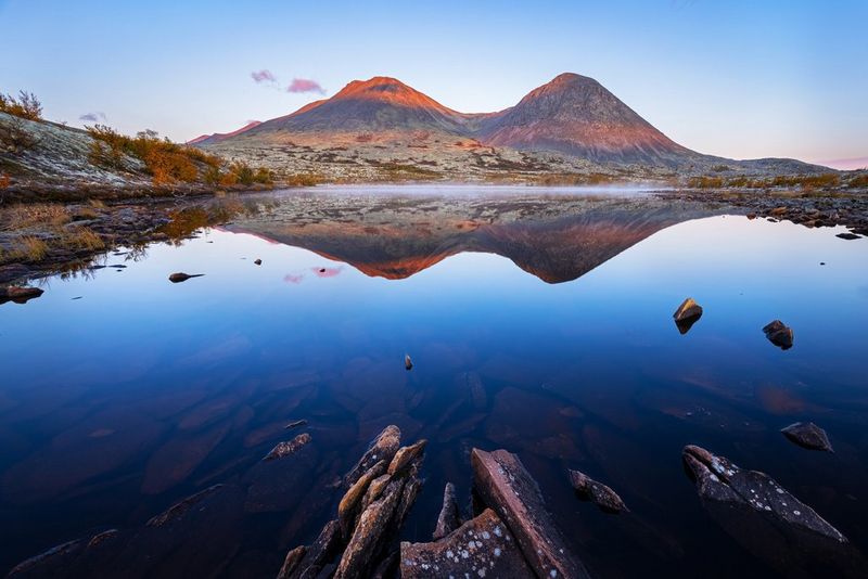 Een bergketen wordt weerspiegeld in een ondiepe plas stilstaand water met ruwe stenen.