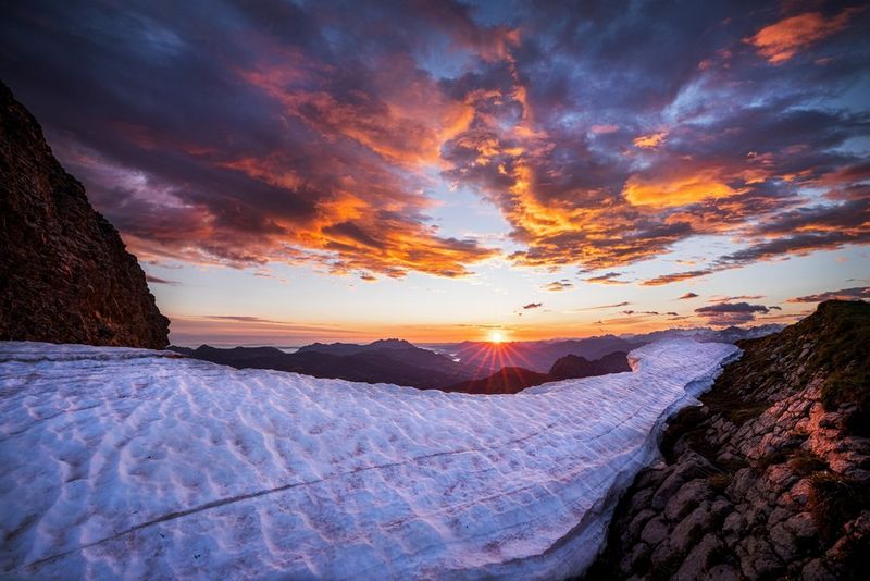 Zachte wolken boven een rotsachtige bergketen met hier en daar bevroren grond, in de oranje gloed van de zonsondergang in de verte.