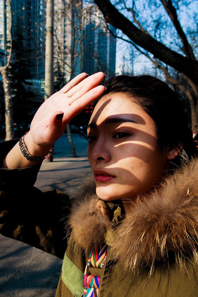 A woman on a city street looks up, blocking the light with her hand, which casts a shadow across her face.