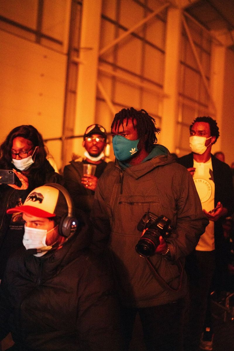 Meji Alabi draagt een mondkapje en houdt een Canon-camera vast op een buitenset, omgeven door zijn filmcrew.