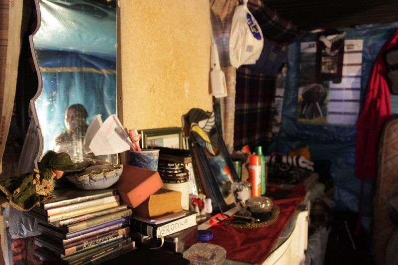 Riflesso di una donna nello specchio di una camera da letto, le sue cose accatastate su un ripiano, in uno scatto di Angela Jimu.