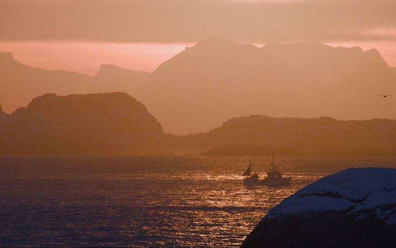 A fishing boat on the glistening water at dusk, with mountains silhouetted behind.