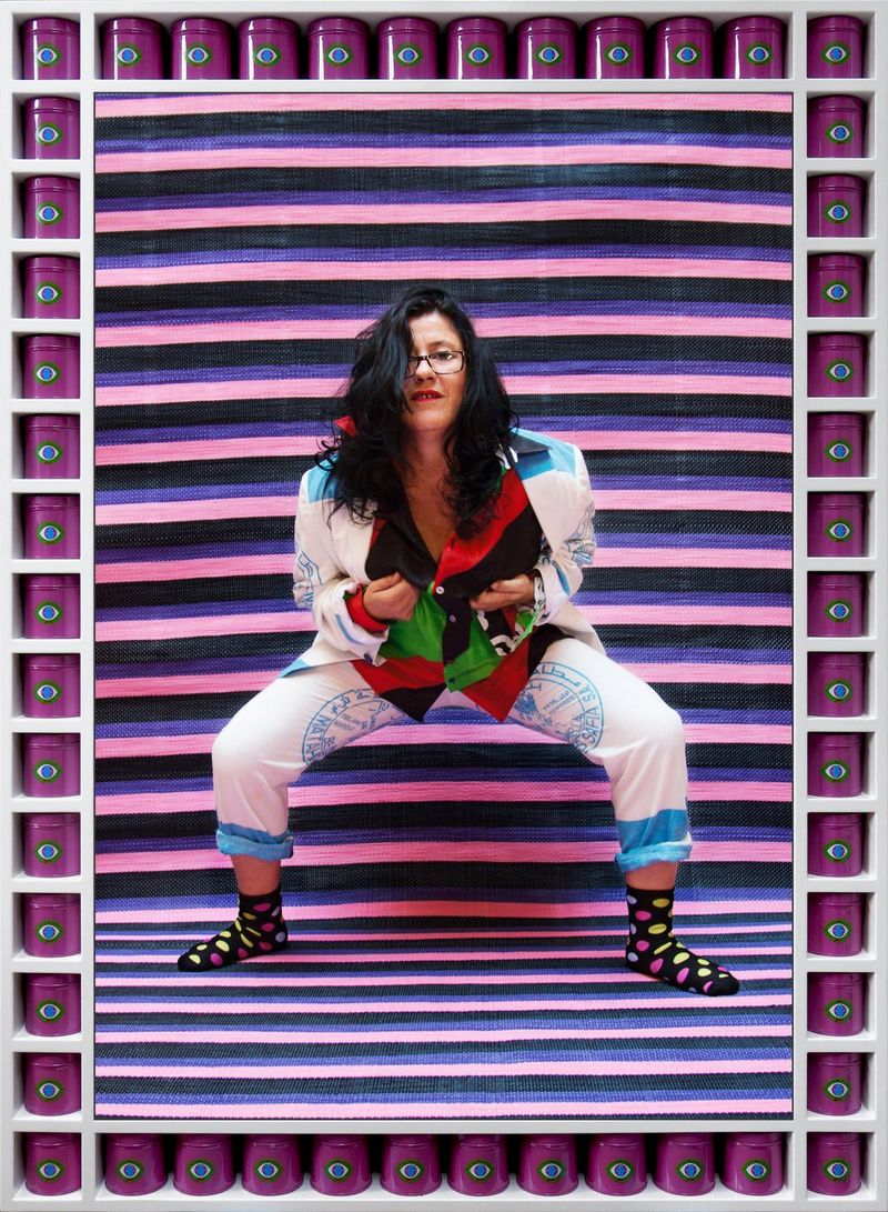 A woman in a white and blue jacket and trousers, with a red, black and green blouse, crouching down in front of a pink, purple and black striped background. The image is framed by purple cans with an eye sticker on each of them. 