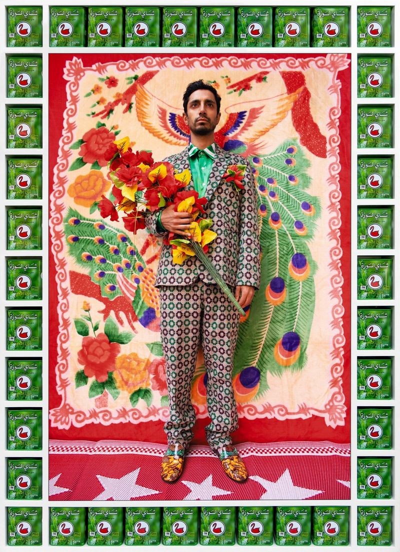A man in a patterned suit holding a bouquet of red and yellow flowers, against a decorative painted background of peacock feathers and flowers. The image's frame is made from green packages. 