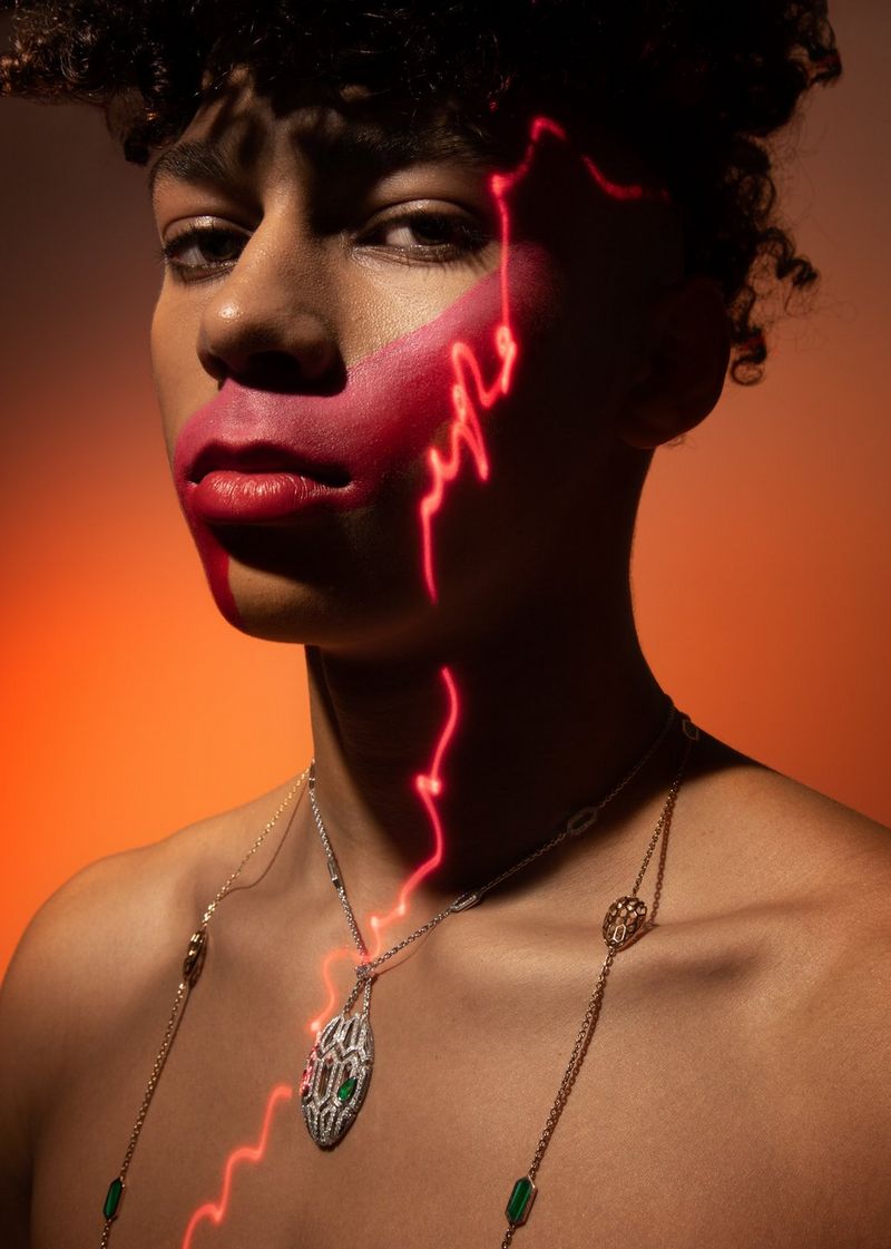 A curly-haired model with a necklace, a red face paint stripe across the mouth and a neon-orange wiggly line running from the eyebrow down the face and neck and across the body.