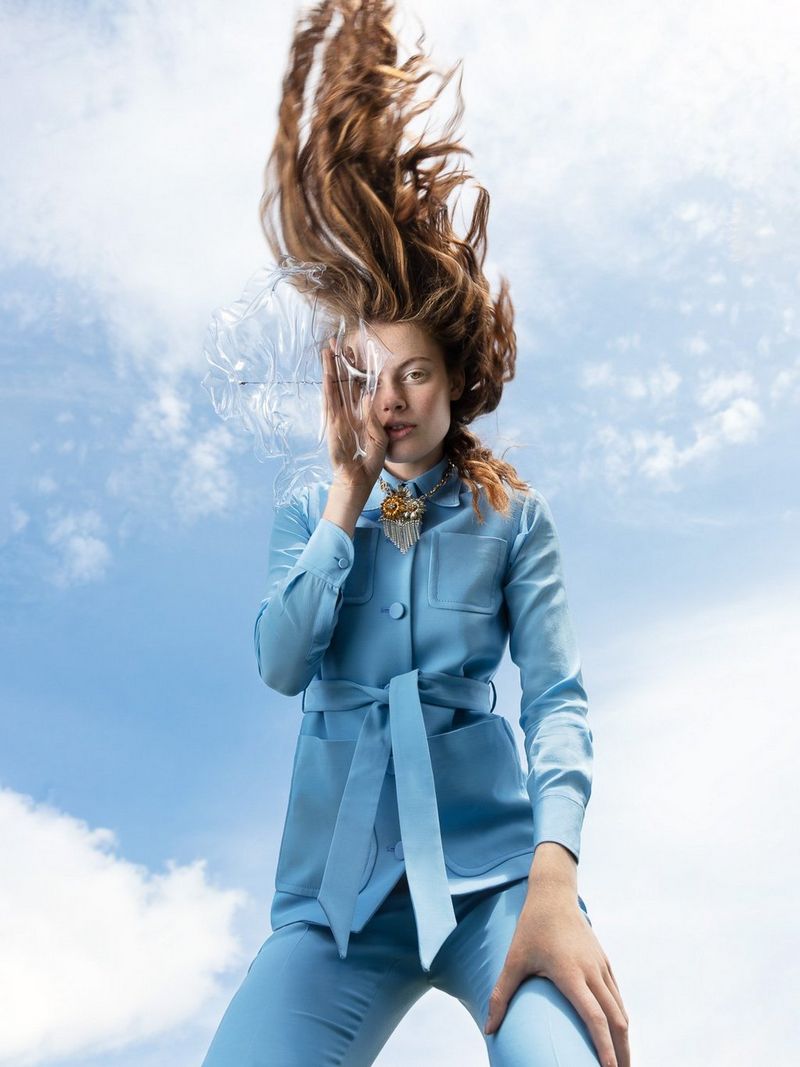 A woman in a blue coat, with the sky behind her, holding a clear plastic object to her face, with her hair appearing to fall upwards into the air. 
