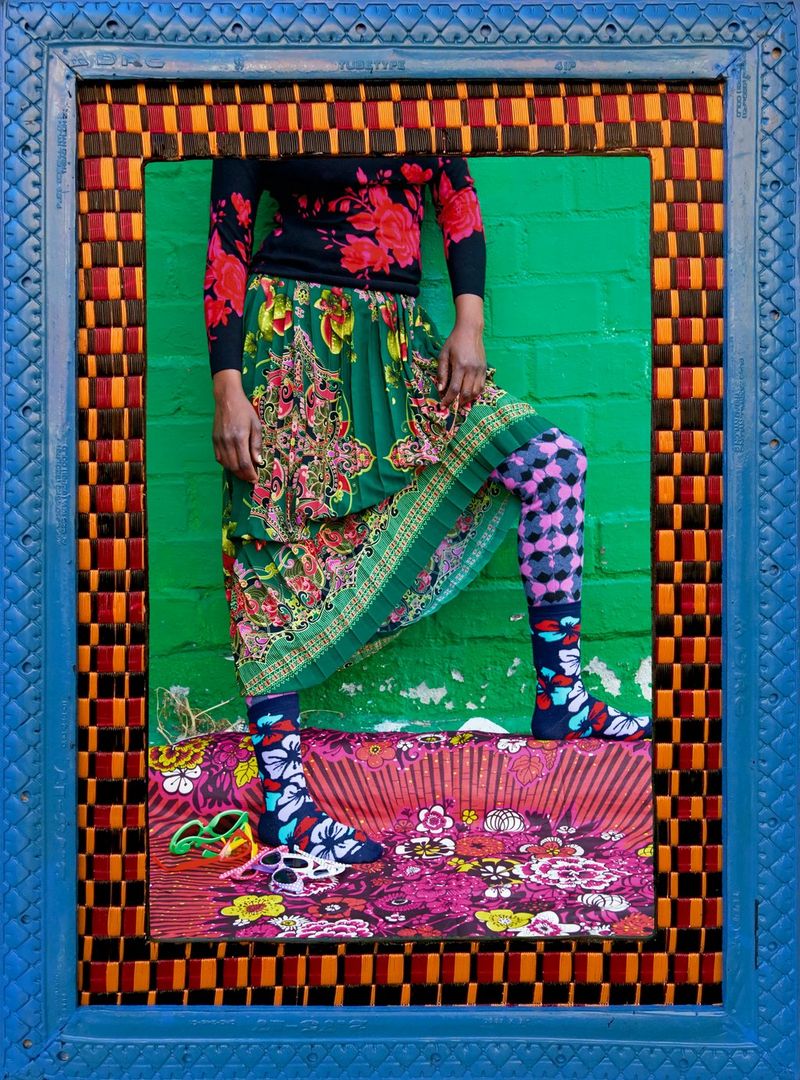 A woman in a bright floral red top and green floral skirt with pink flowers, with her shoulders and head out of frame, in front of a green brick wall. The image has two frames, one blue with a scale pattern, the other appearing to have been weaved like a basket.