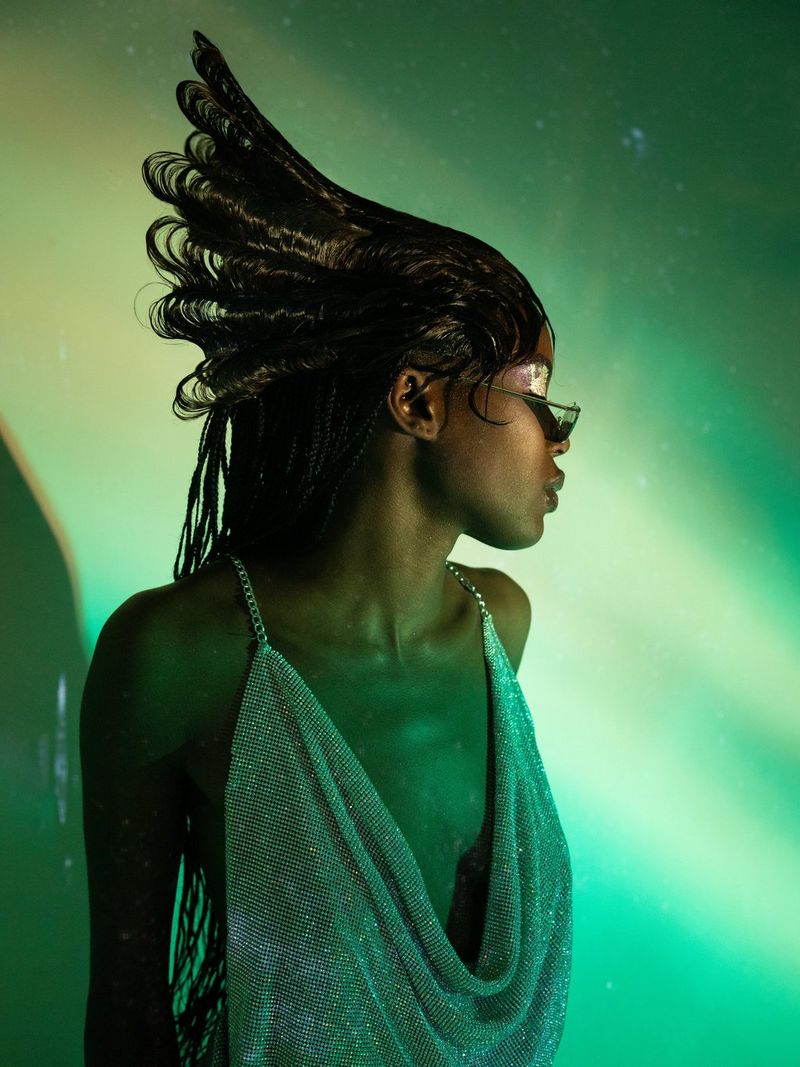 A woman with an elaborate hairstyle flared behind her head, wearing a deep-cut, shimmering top, with green light shining on her. 