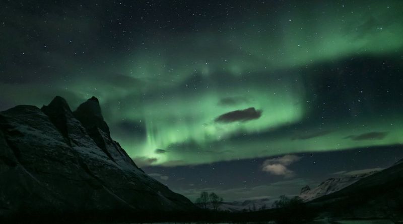 The skies above a mountain peak lit with green light trails from an aurora.