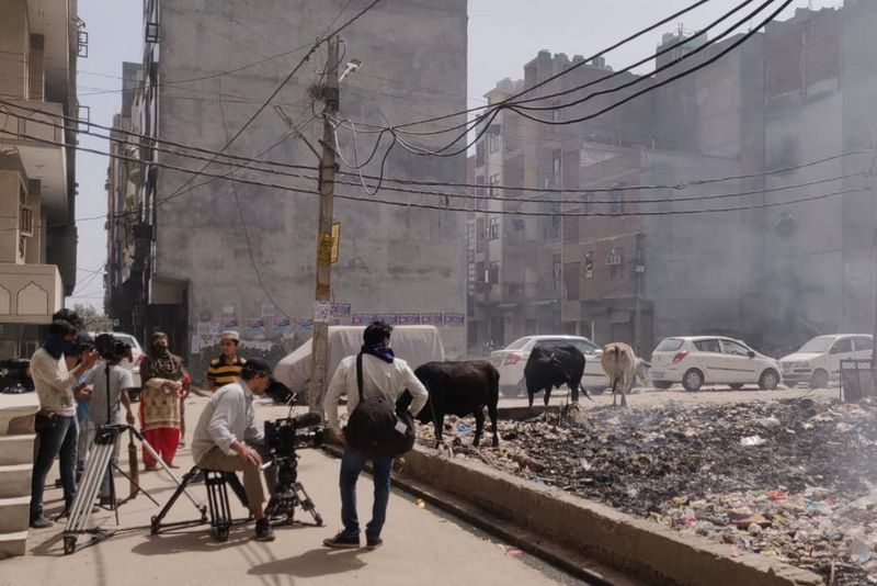 A film crew recording in the streets of Delhi, among cars and cows, and next to a smouldering pit of rubbish.