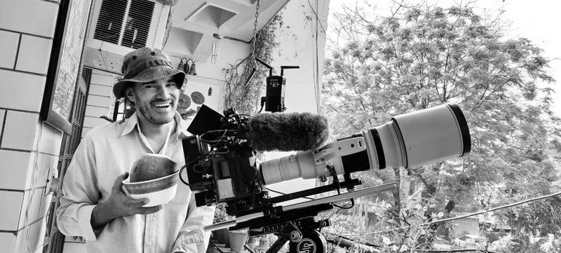A man stands smiling on a balcony next to a film camera setup in this black and white image.