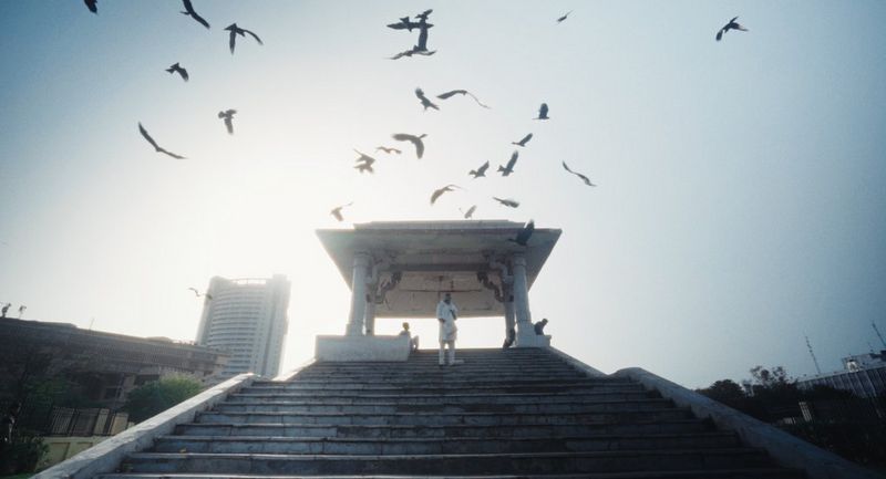 Figures stand at the top of steps with birds swooping all around against the sky.