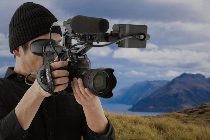 A man stands on hilltop holding a Canon EOS C500 Mark II. Mountains can be seen in the background.