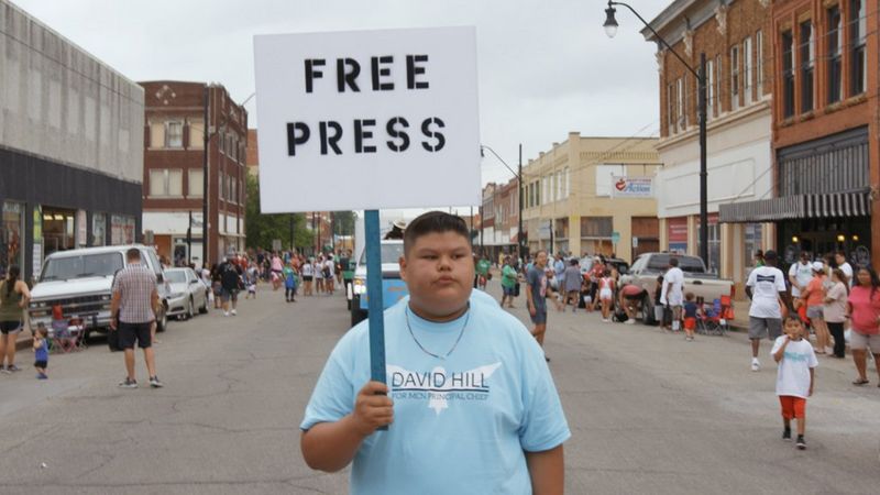 A still from the documentary Bad Press, filmed on a Canon EOS C300 Mark II, showing a young man in a busy street holding a placard with the words 'FREE PRESS' on it.