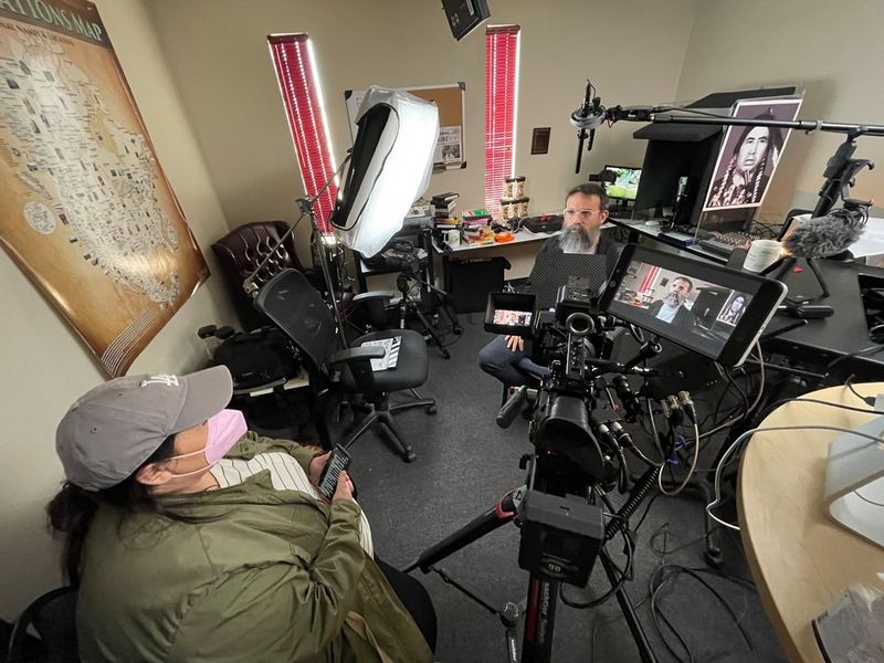 A man with a long grey beard sits in the middle of a cluttered office surrounded by filming equipment, including a Canon EOS C300 Mark II. A second person sits on a chair out of shot facing him.