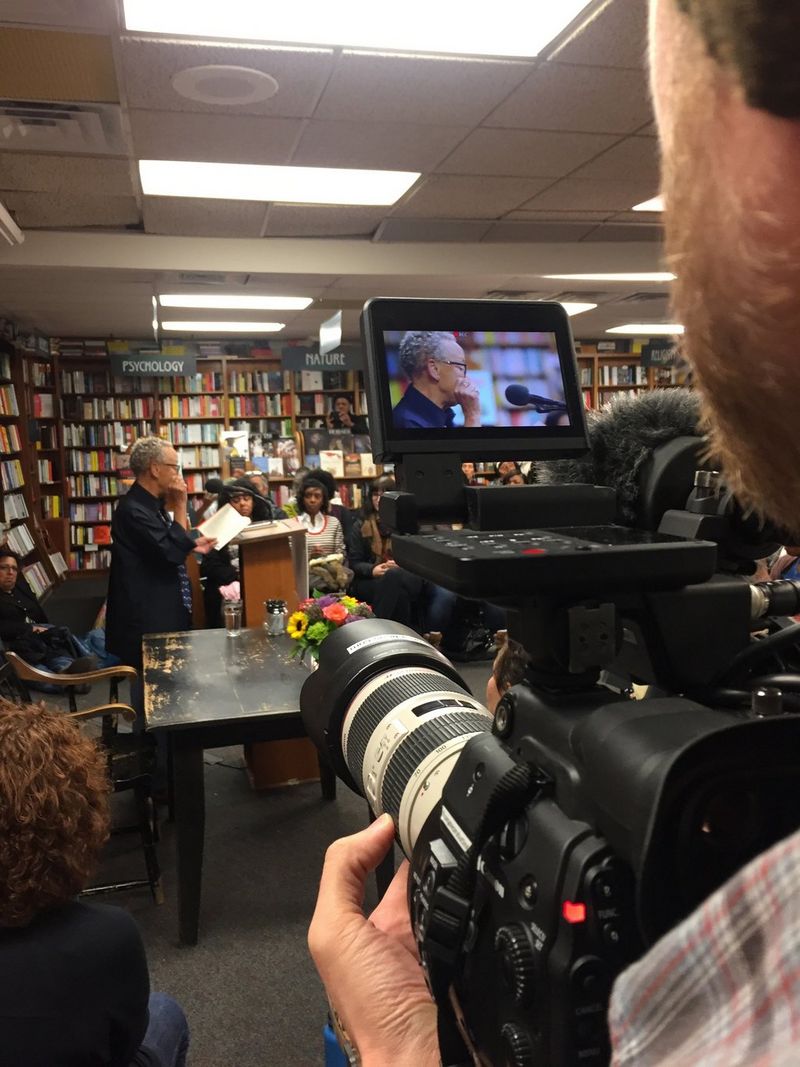 A woman stands at a lectern in a bookshop reading from a book in front of her. She is being filmed by a man standing at the front of the shot, using a Canon EOS C300 Mark II on a tripod.