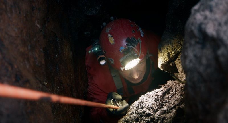 A film still of male miner abseiling down a mine in a very tight space, with a head torch on his hard hat lighting the stone in front of him, captured on the Canon EOS C70.