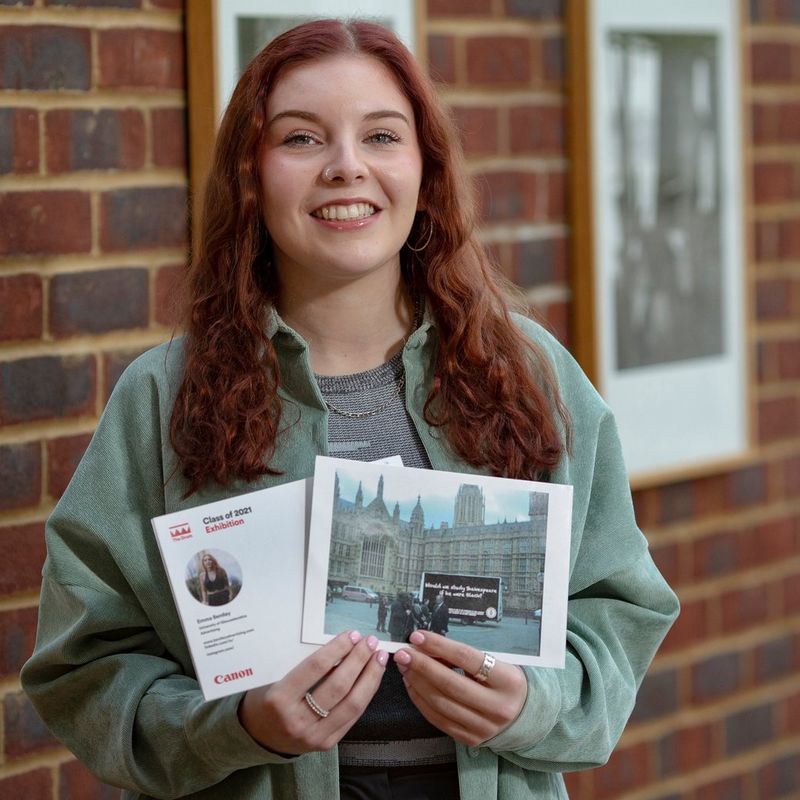 A young woman holds up printed A5 postcards featuring her profile and examples of her work. Behind her is a brick wall covered in framed black and white prints.