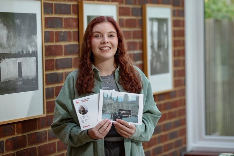 Een jonge vrouw houdt geprinte A5-briefkaarten vast waarop haar profiel en voorbeelden van haar werk te zien zijn. Ze staat voor een stenen muur met ingelijste zwart-witte prints.