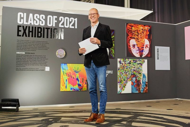 Presentator en verslaggever Richard Askam staat voor een tentoonstellingswand, waarop enkele kunstwerken en tekst te zien zijn.