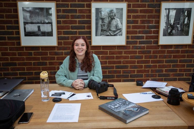 Een jonge vrouw zit achter een bureau met een Canon EOS R6 naast haar en met ingelijste, zwart-witte foto's op de muur achter haar.