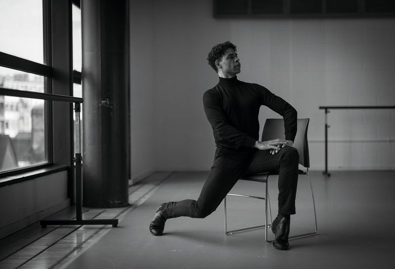 Een zwart-witportret van een man in een zwart shirt en zwarte broek die op een stoel voor een raam poseert. Hij leunt voorwaarts op zijn tenen en zijn handen rusten op zijn been.
