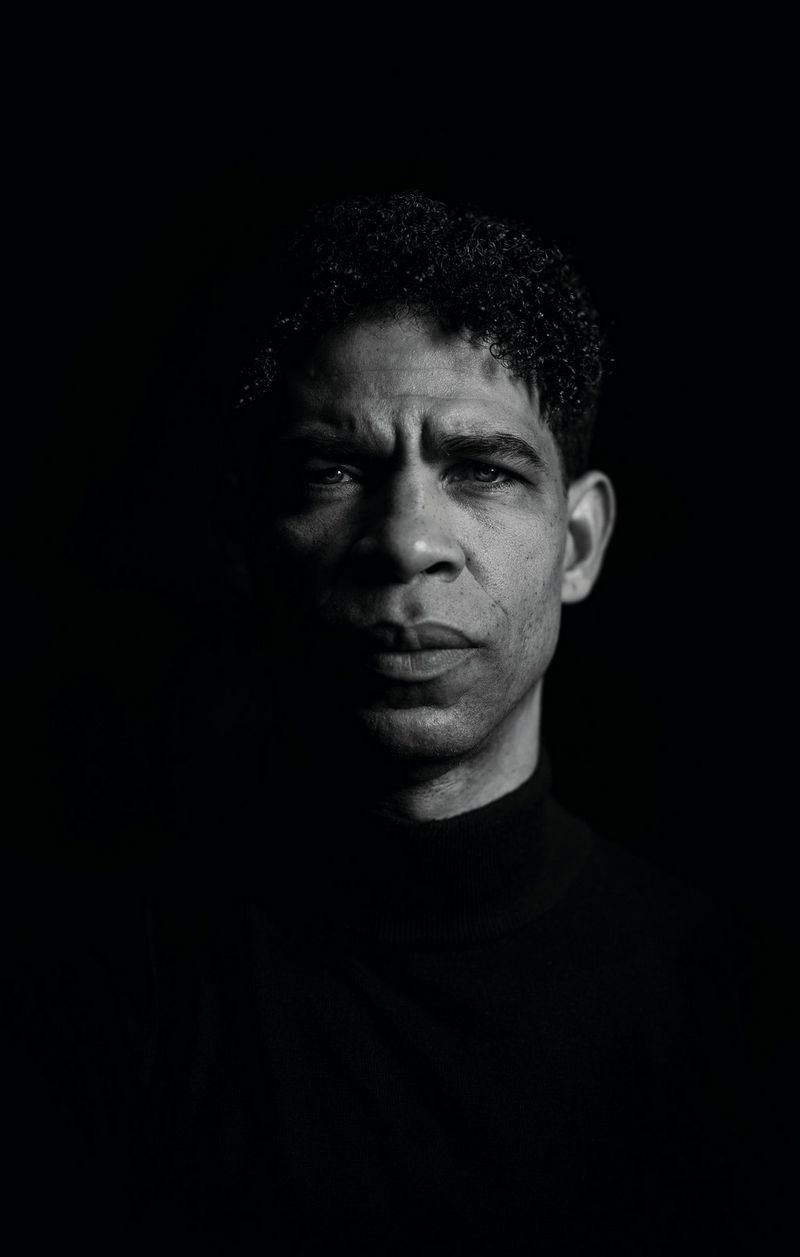 Een zwart-witportret van het gezicht en de schouders van een man. Zijn haar en shirt zijn zwart, en gaan op in de zwarte achtergrond.