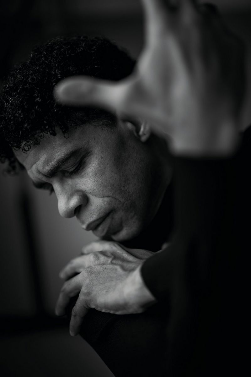 Een zwart-witportret van een man die zijn armen gekruist voor zich houdt. Een hand ligt op zijn schouder en de andere houdt hij in de lucht voor zich, dicht bij de camera.