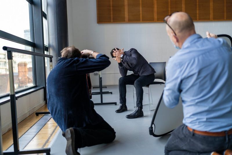 Een fotograaf knielt om een foto te nemen van een man op een stoel met beide handen voor zijn gezicht. Een andere man knielt ernaast en houdt een groot reflectiescherm vast.