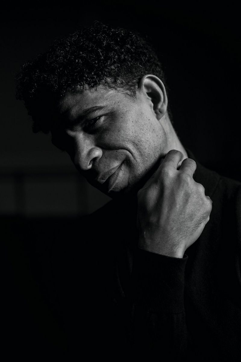 Een zwart-witportret van het gezicht en de schouders van een man die een hand bij zijn nek houdt. Zijn haar en shirt zijn zwart, en gaan op in de zwarte achtergrond.