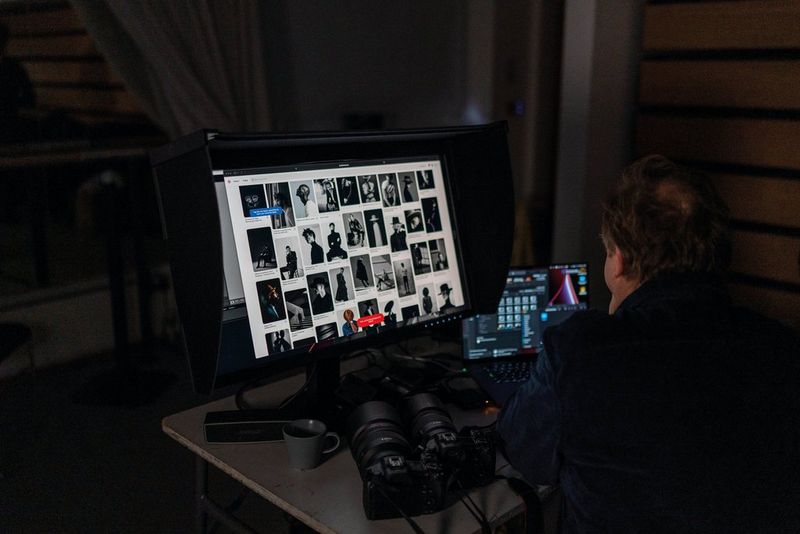 Een man zit aan een bureau naast twee camera's. Hij werkt op een laptop en met een grote monitor waarop een uitgebreide collectie portretfoto's wordt weergegeven.