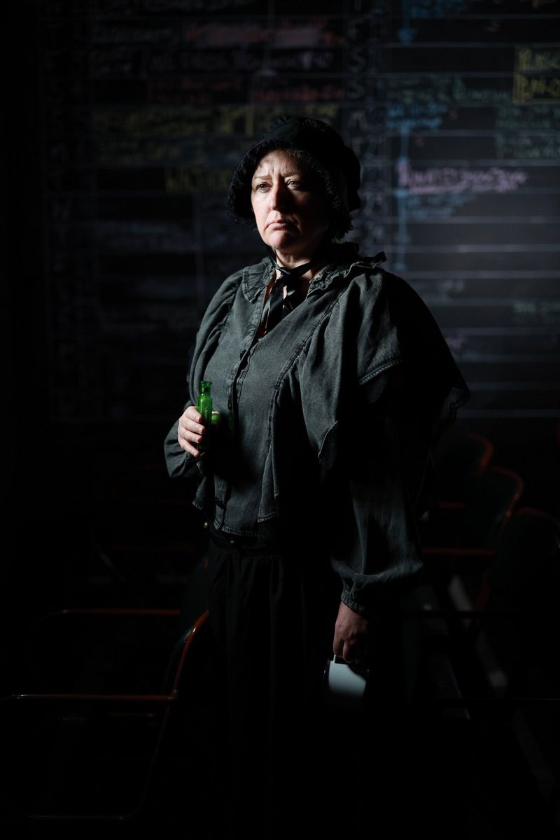  A portrait of a woman wearing a green tunic and a black bonnet, and holding a green glass bottle, photographed by Clive Booth.