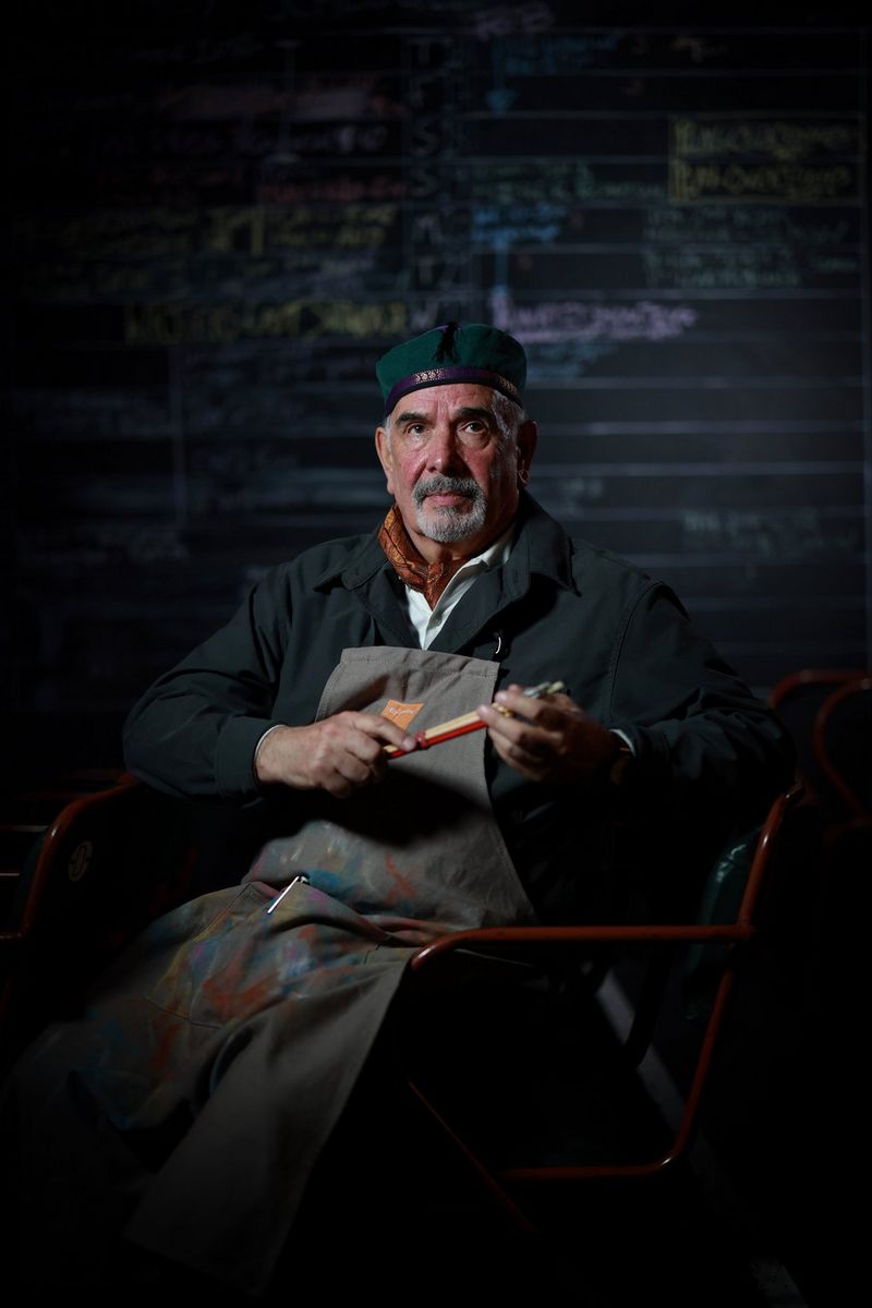 A portrait of a man wearing an apron covered in paint stains, holding paintbrushes in his hand while sitting in a chair, photographed by Clive Booth. 