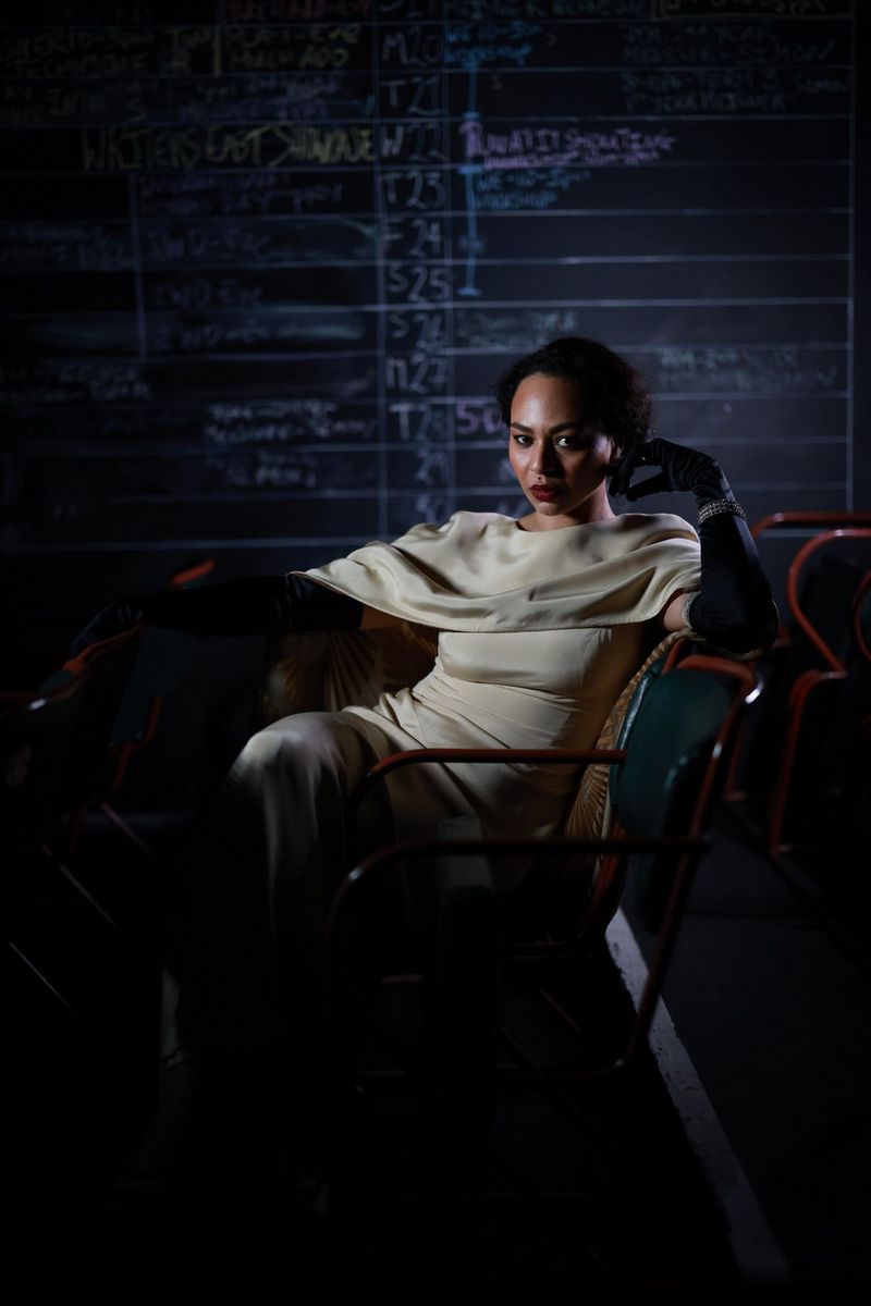 A portrait of a woman wearing a cream satin dress and long black gloves while sitting in a chair, photographed by Clive Booth. 