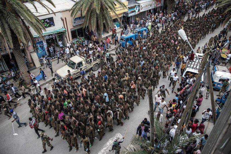 An overhead shot of hundreds of soldiers marching through the streets flanked by men carrying batons.