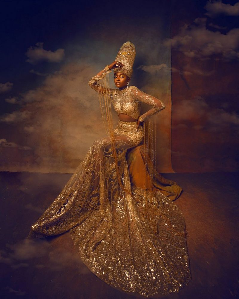 Una donna seduta con indosso un top e una gonna di perline dorati molto elaborati e un copricapo alto. Lo sfondo raffigura un cielo con nuvole. Scatto realizzato da Emmanuel Oyeleke con obiettivo Canon EF 16-35mm f/2.8 L III USM.