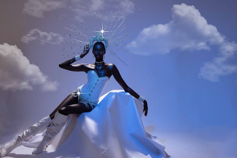Una donna con indosso un corsetto bianco, stivali alti al ginocchio e un elaborato copricapo luccicante è appoggiata a un lenzuolo bianco drappeggiato, con sullo sfondo un cielo blu-viola e soffici nuvole bianche. Scatto realizzato da Emmanuel Oyeleke con Canon EOS R5.