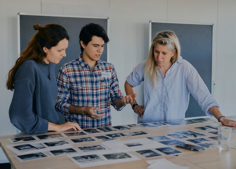 Photojournalist Ahmed Qabel stands at a table between two women, looking at a large selection of photographs spread out in front of them.