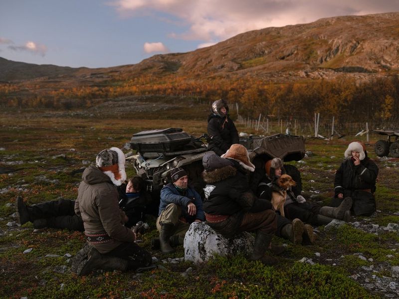 A group of people in warm hats and clothing sit on the ground resting, with hills in the background, in a photograph by Michał Siarek