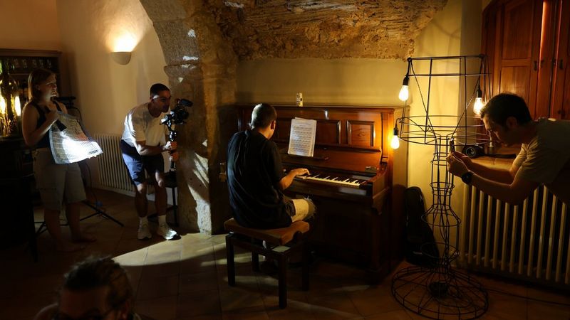 A photographer sets up a shot of a man playing a piano in low light, with another person shining a torch on a reflector and a third person taking a shot with a smartphone. 