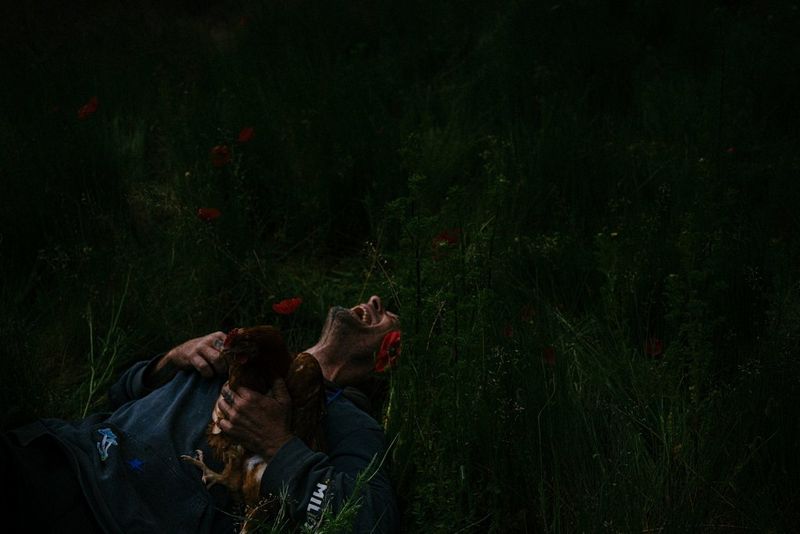 A man lying in long grass, laughing, head thrown back, holding a chicken to his chest. Photo by Emeline Sauser 