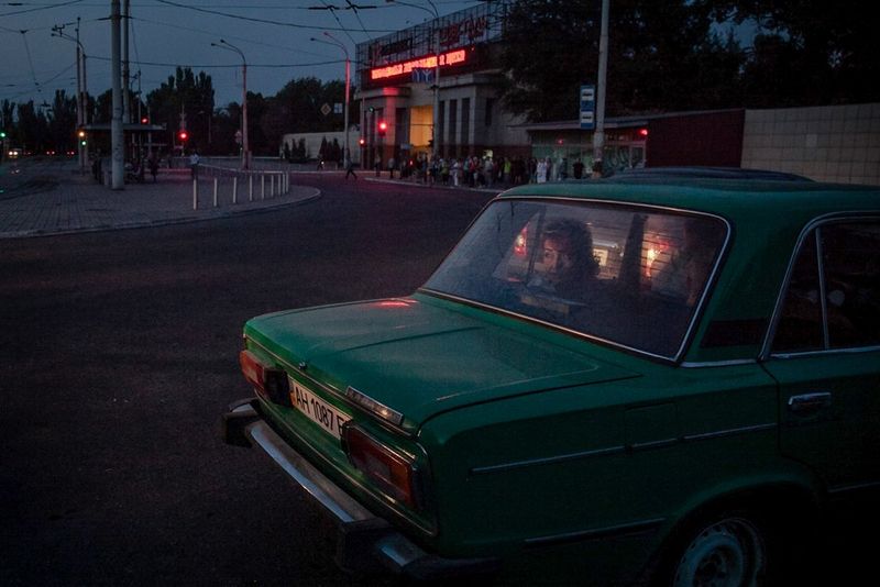 A green car is parked near a quiet intersection at dusk, with two passengers visible inside looking out the back window. In the background, a building with neon signage and a small crowd of people can be seen. Photo by Simona Supino.