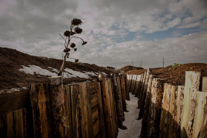 A desolate wooden trench stretches into the distance under a cloudy sky. Snow patches and a lone plant are visible on the dirt embankments lining the trench. Photo by Simona Supino
