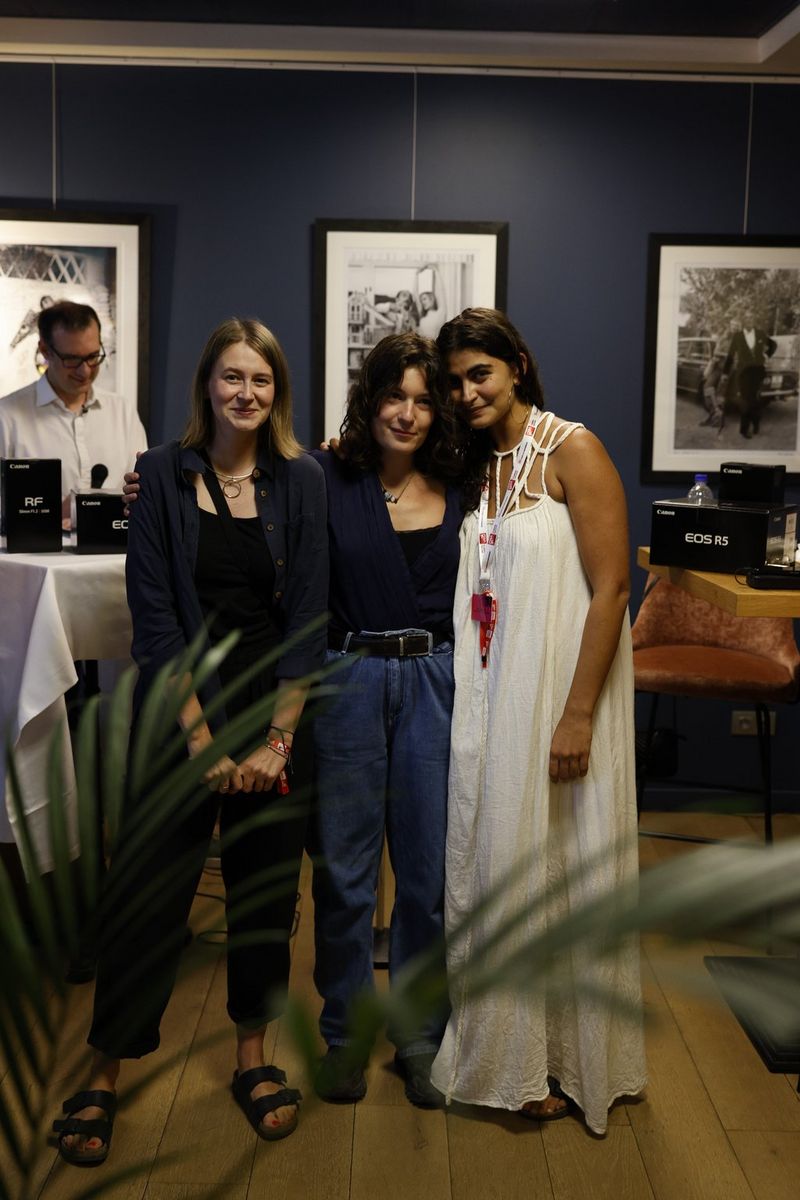 Three women stand together posing for a photo in a room with framed pictures on the wall behind them.