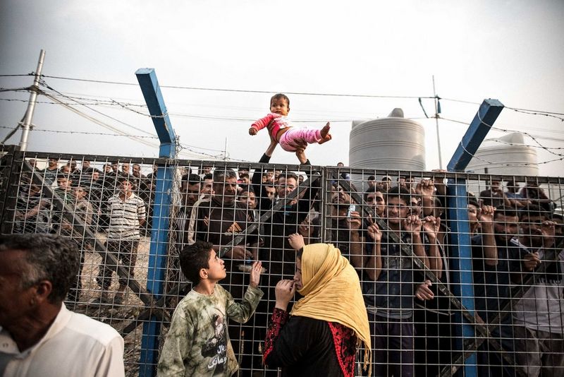 A photo by Daniel Bateman showing a small child being held aloft over a fence topped with barbed wire, with a crowd of figures looking through from behind.