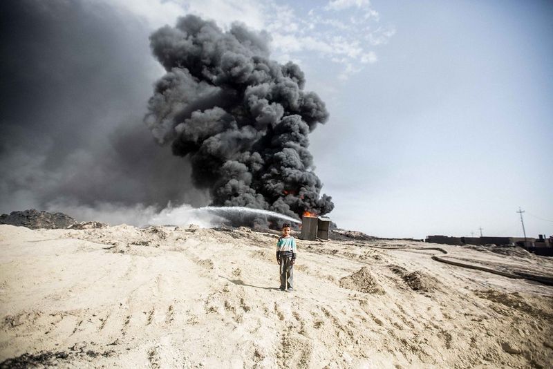 A photo taken by Daniel Bateman showing a small boy standing in a desert in Iraq with black smoke from a fire billowing into the air in the background. 