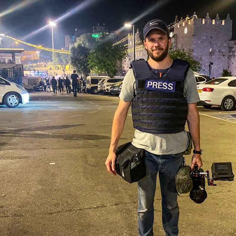 Daniel Bateman stands on a street holding his Canon Cinema EOS camera in one hand. Behind him, people are walking towards a crowd apparently gathering in the background.