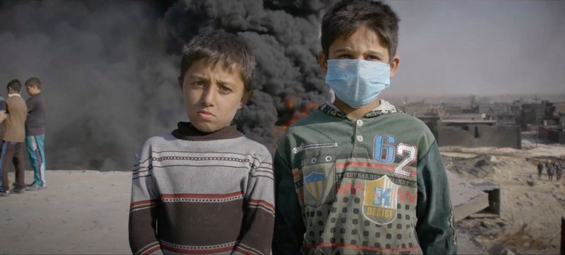  Two children, one wearing a facemask, stand in front of a fire and plume of smoke.