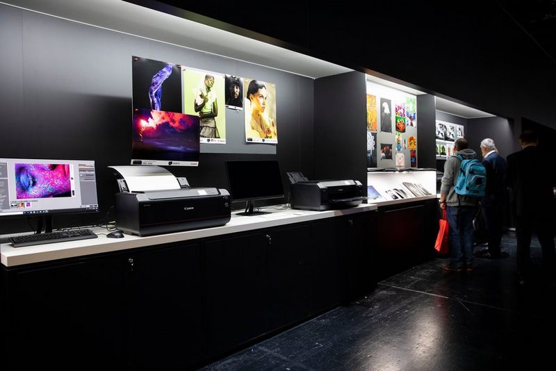 A Canon display stand featuring two Canon PIXMA PRO-100 printers, a series of colourful prints and a monitor with a woman's highly stylised face on it. 