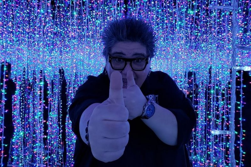Deborah Corn gives two thumbs-up to the camera. She is in a dark room lit by many strands of multi-coloured fairy lights.